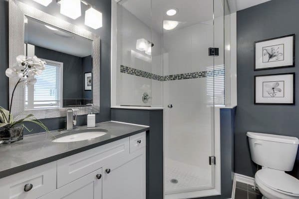 Modern bathroom with a corner glass shower, dark countertops, white cabinets, and framed art on gray-blue walls