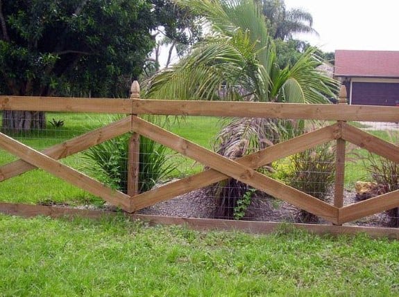 A wooden zigzag fence with wire mesh stands in front of green grass, palm trees, and a house in the background