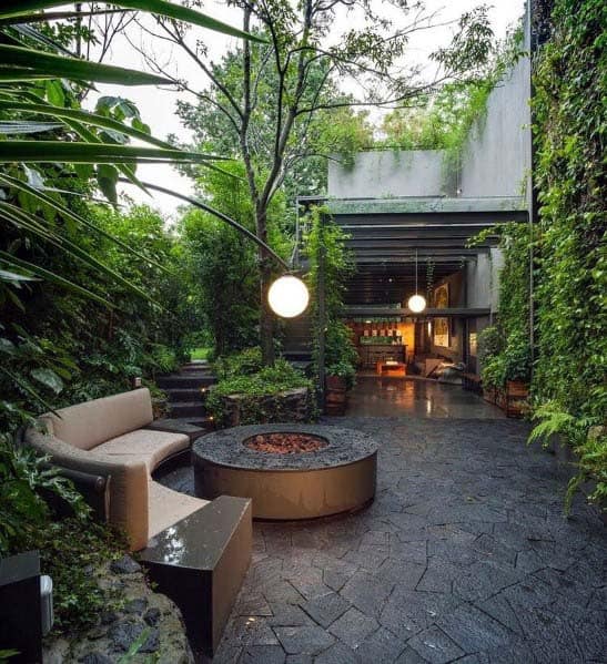 Modern outdoor patio with a round fire pit, cozy seating, lush greenery, and hanging lights