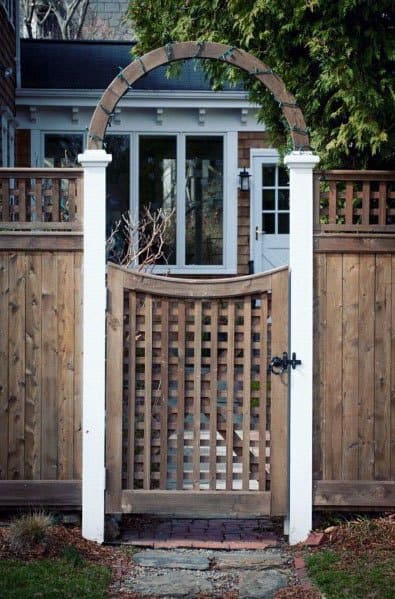 Wooden garden gate with an arch and lattice design, flanked by tall picket fences, leading to a house