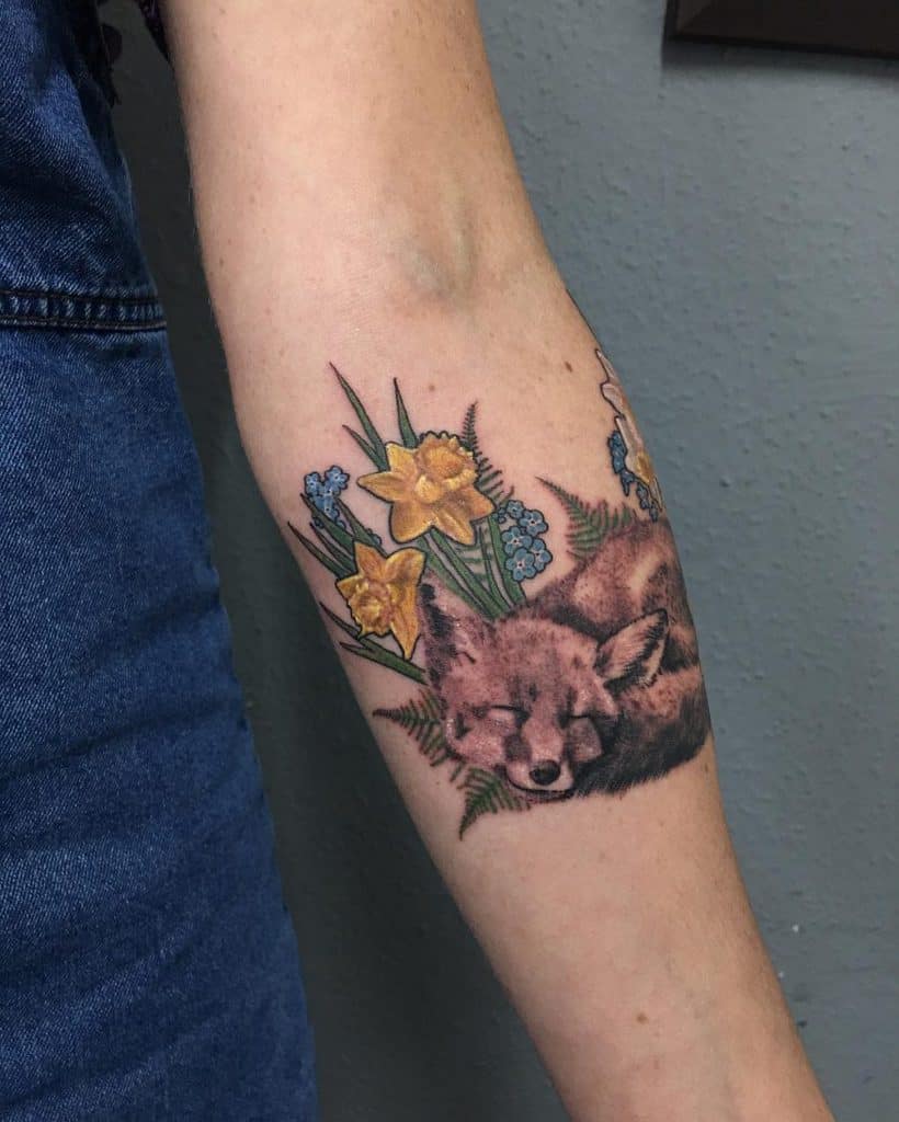 A peaceful tattoo of a sleeping fox surrounded by vibrant daffodils and colorful wildflowers, combining nature’s beauty with a serene atmosphere.