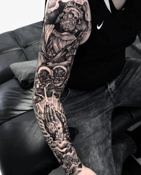 Black and grey full-sleeve tattoo featuring a detailed depiction of a crowned Jesus, cherubs, praying hands, and intricate shading for a religious theme.
