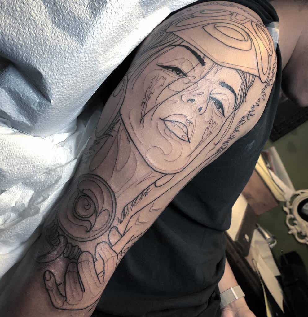 A line art tattoo of a character wearing a headdress, showing a close-up of the face with facial markings. The design includes intricate detailing and outlines of the character's facial features and hand.