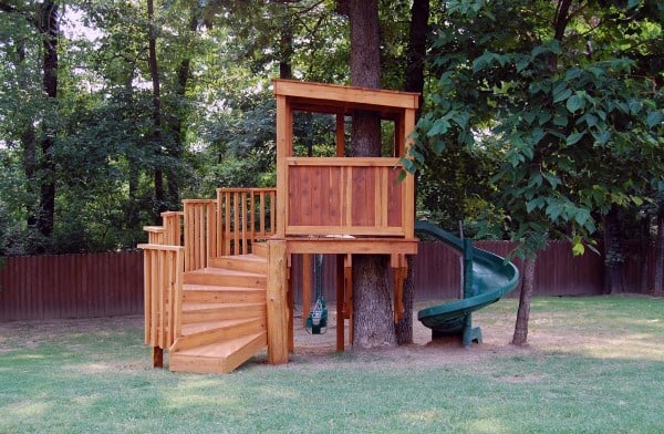 Wooden treehouse with stairs and a green spiral slide in a grassy backyard with trees