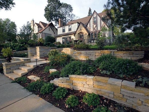 Elegant front yard slope landscaping with tiered stone retaining walls, lush greenery, and steps leading up to a Tudor-style home
