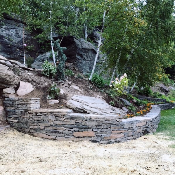 A stacked stone retaining wall on a sloped garden, creating a terraced effect with plants and flowers along the edge