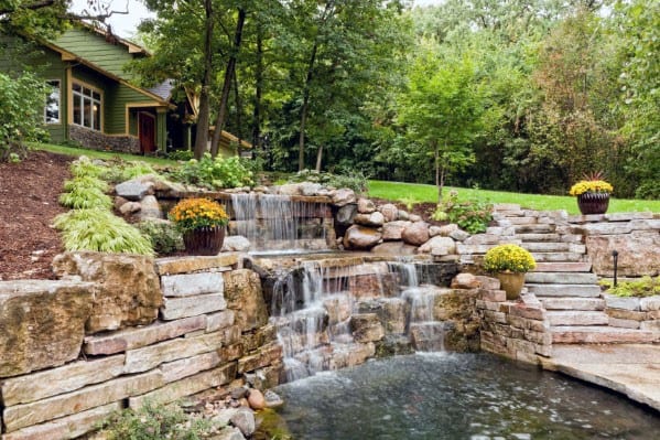 Sloped landscape featuring a multi-tiered stone waterfall, tranquil pond, stone steps, and lush greenery for a serene garden retreat