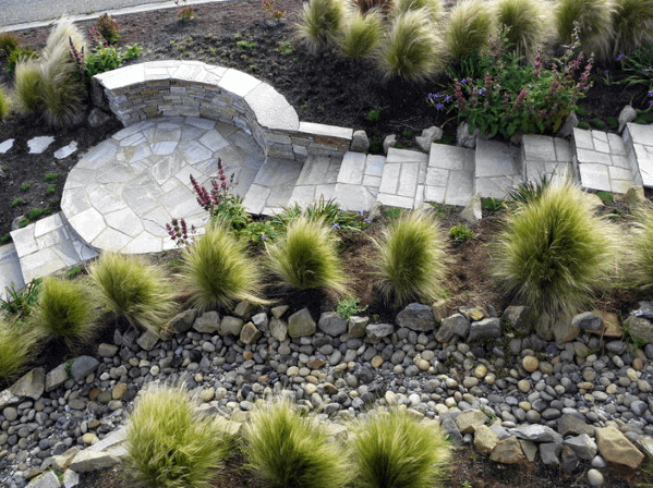 Elegant stone walkway with river rock landscaping, leading to a circular stone seating area, surrounded by lush grasses and plants