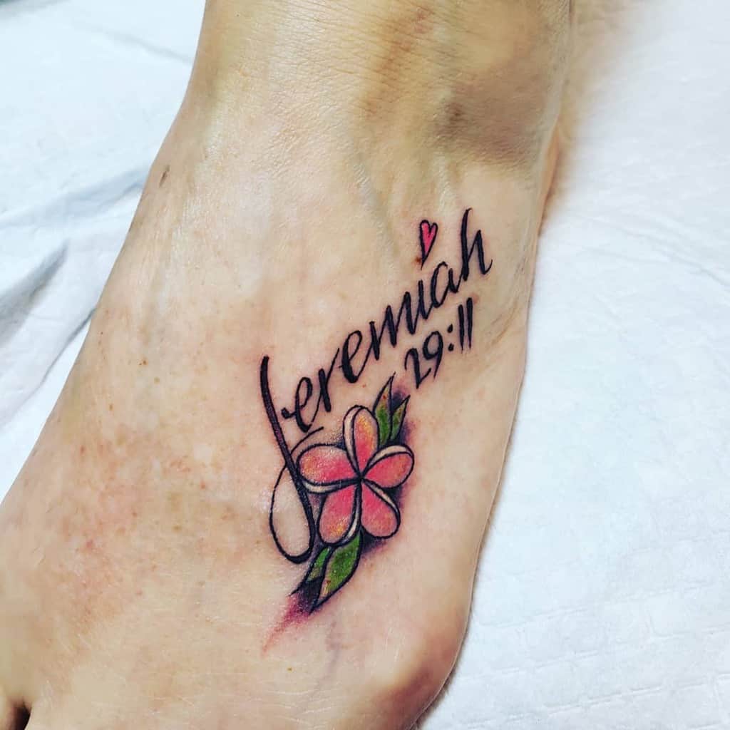 Foot tattoo featuring "Jeremiah 29:11" alongside a vibrant red plumeria and lush green leaves