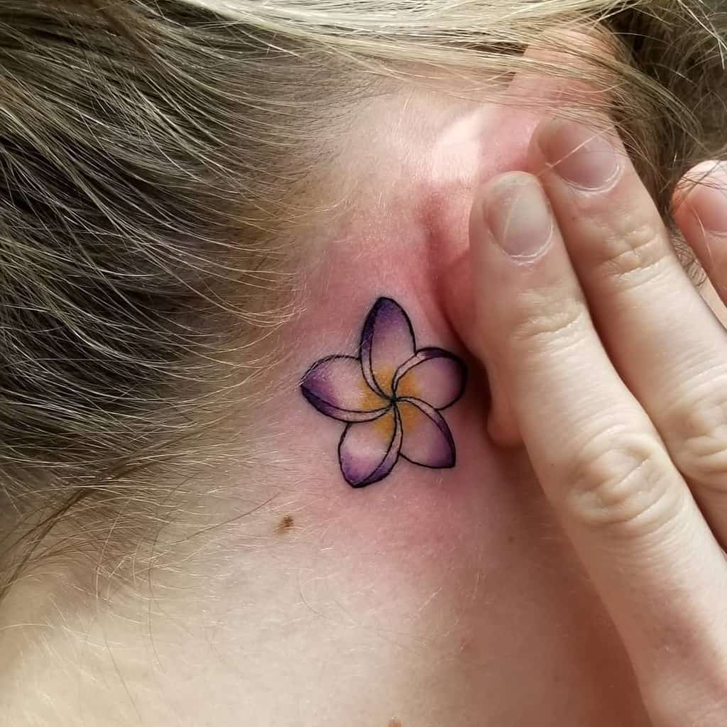 A delicate plumeria flower tattoo graces the area behind the ear, beautifully shaded in hues of purple and yellow