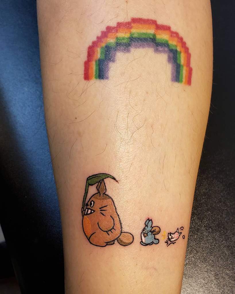 A playful Totoro tattoo featuring a small Totoro holding a leaf as an umbrella, accompanied by friends under a pixelated rainbow