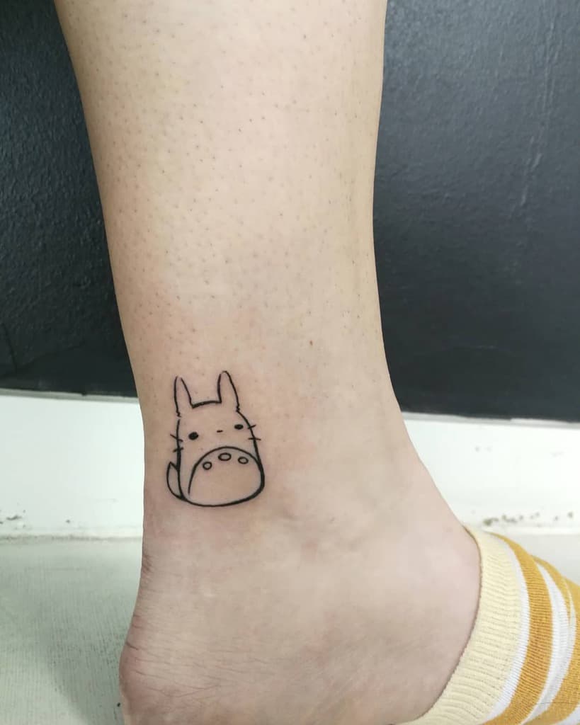 Simple and minimalist Totoro tattoo on the inner ankle, featuring Totoro’s iconic face in black line art