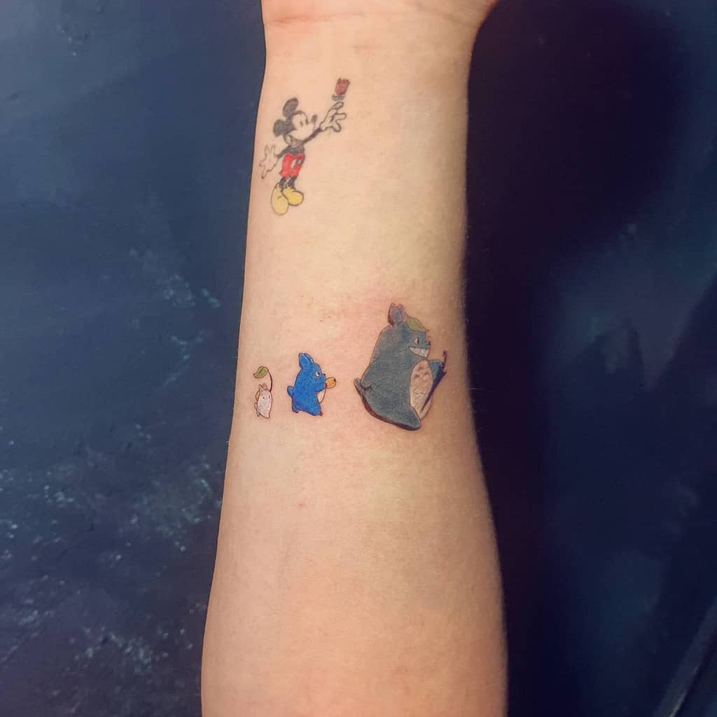 A minimalist Totoro tattoo design featuring three small Totoro characters in a walking sequence, placed subtly on the forearm, adding a touch of whimsy