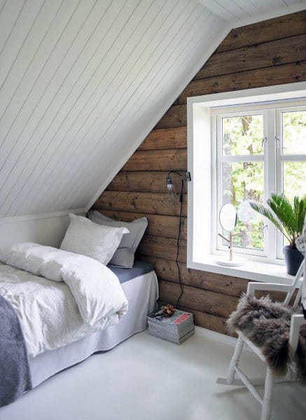 Cozy attic bedroom with wooden walls, a white ceiling, a bed, and a rocking chair by a window overlooking trees
