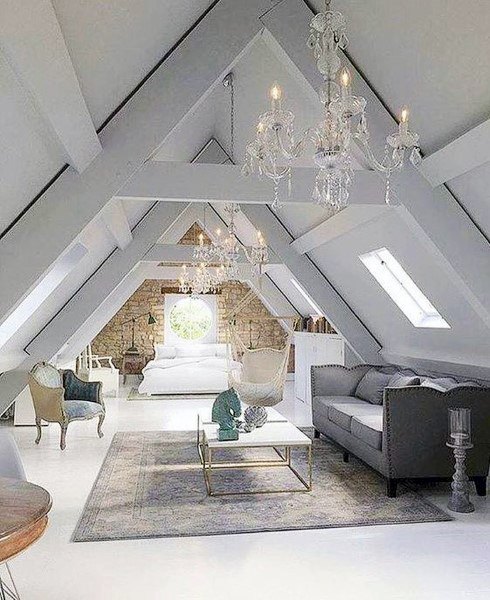 A stylish attic room with chandeliers, sofa, armchairs, bed, sloped ceiling, and circular window