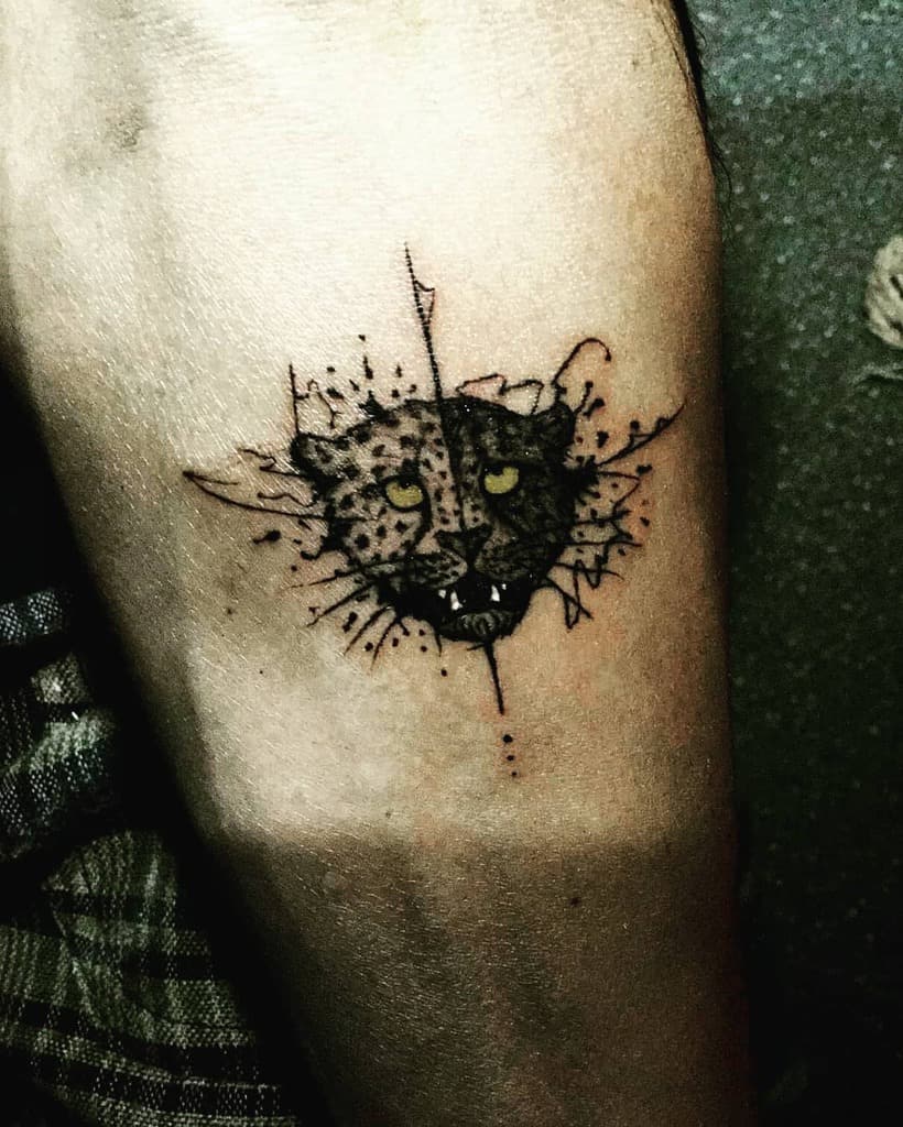 A dynamic black ink tattoo of a jaguar's face emerging from a splatter effect, with bold details and fierce eyes, placed on the forearm.