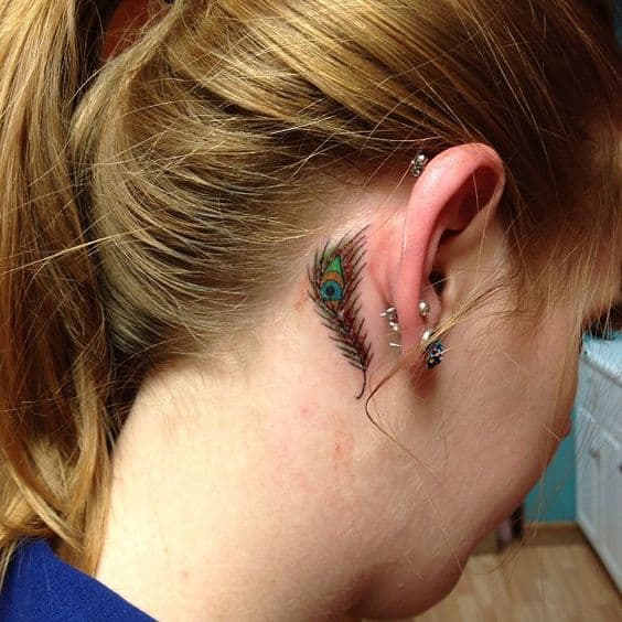 Small peacock feather tattoo behind the ear, featuring delicate green, blue, and gold hues with fine detailing for a subtle and elegant look.