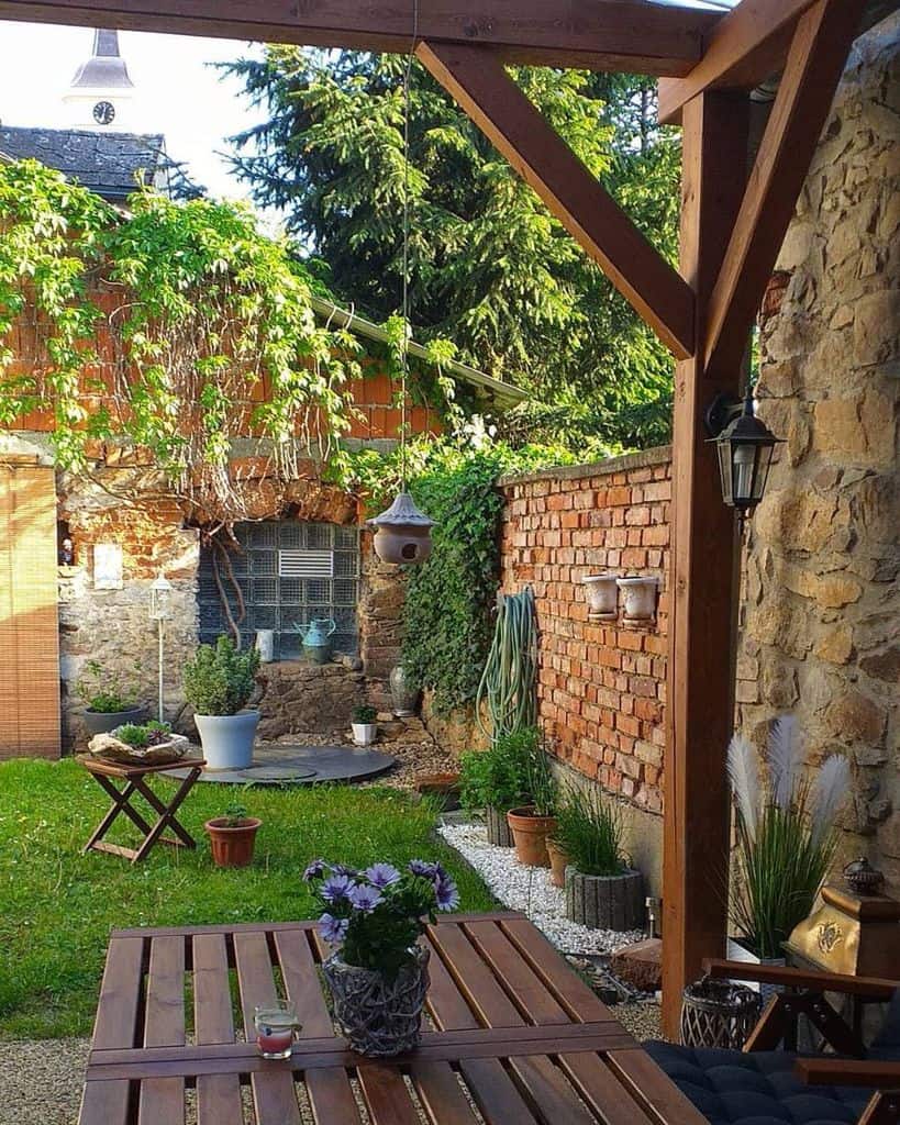Cozy backyard patio with wooden pergola, table, hanging birdhouse, brick wall, plants, and garden view