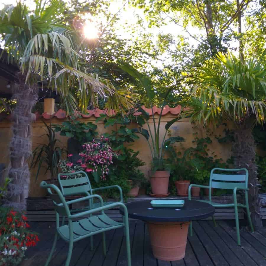 Garden patio with green chairs, a round table, and potted plants. Sunlight filters through lush trees and tropical foliage