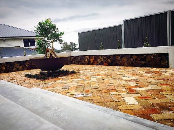Brick patio area with a cozy fire pit, stacked wood, low white wall, tall fence under an overcast sky