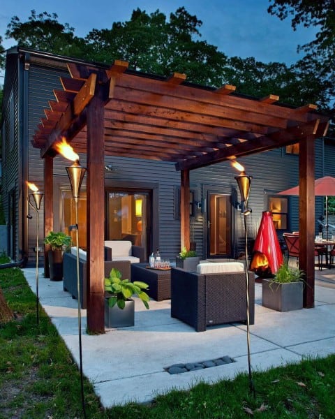Modern patio features a wooden pergola with seating, torches, and lush plants at dusk