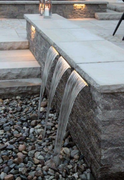 Elegant stone waterfall feature with three streams flowing over smooth pebbles in a patio setting.