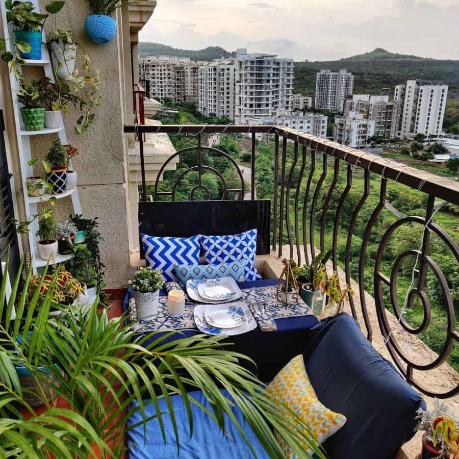 Cozy balcony with blue cushions, potted plants, and dining setup overlooking a cityscape.