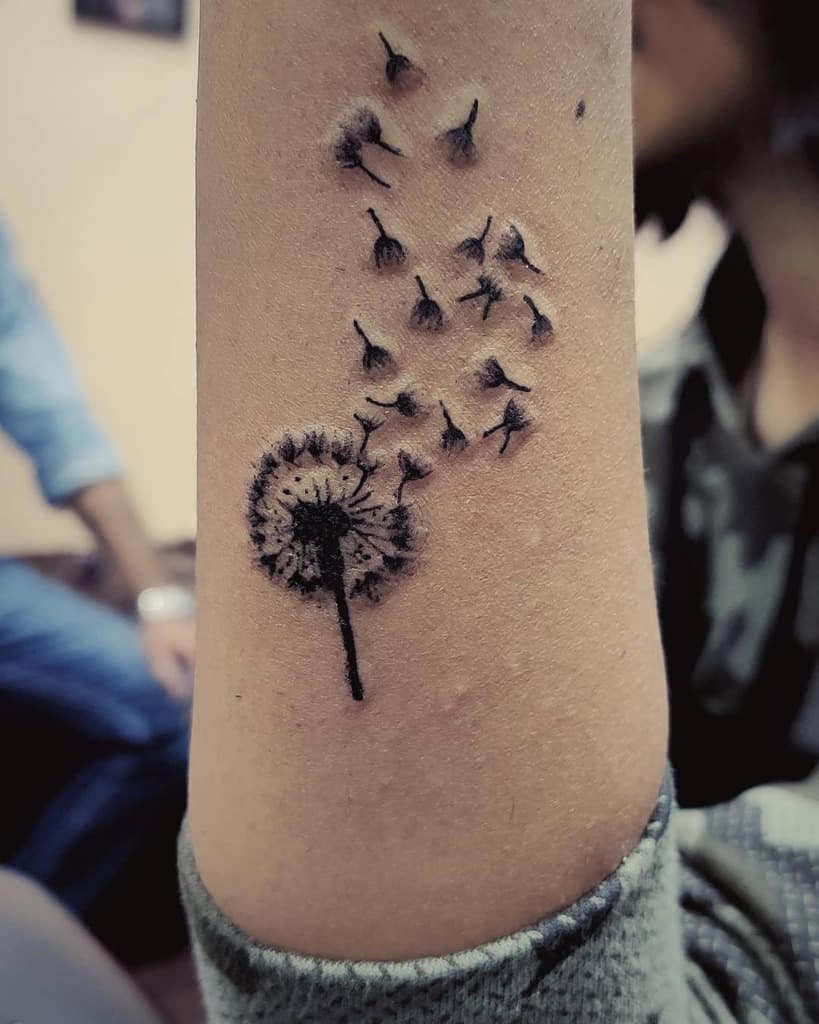This tattoo features a dandelion with seeds blowing away, symbolizing movement and change. The design captures both delicacy and strength, placed on the arm for a flowing, dynamic effect.