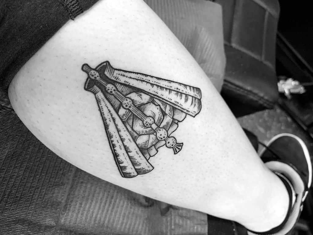 Black ink tattoo on the thigh featuring a detailed hand gripping three wands, with bold linework and shading for a vintage engraving effect.