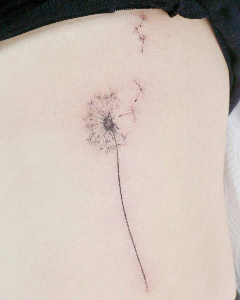 Fine-line dandelion tattoo on the ribcage with seeds floating away, symbolizing freedom, dreams, and life’s fleeting moments.
