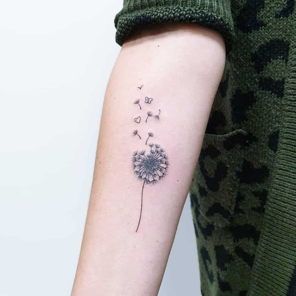 This tattoo features a dandelion with seeds dispersing into the air, accompanied by butterflies and hearts. It conveys a sense of lightness, freedom, and love, beautifully done in fine lines. The design is delicate and meaningful, making it a captivating piece.