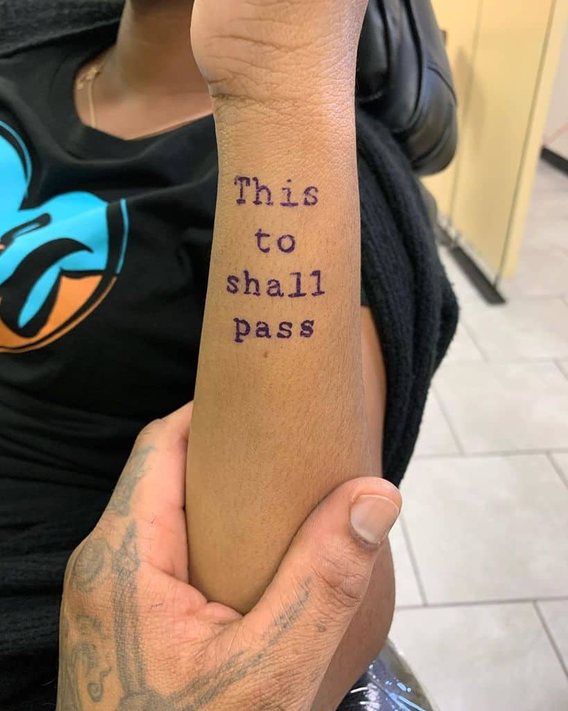 The image shows a tattoo of the phrase "This too shall pass" on the person's arm.