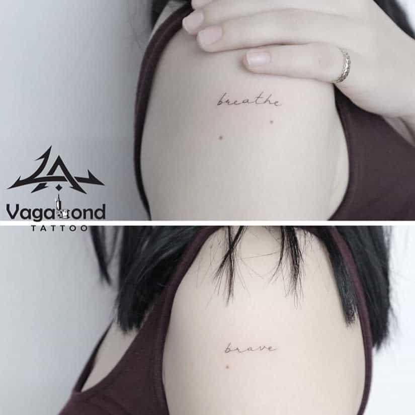 Minimalist tattoos on the shoulder with the words 'breathe' and 'brave' in delicate cursive font, inked in black