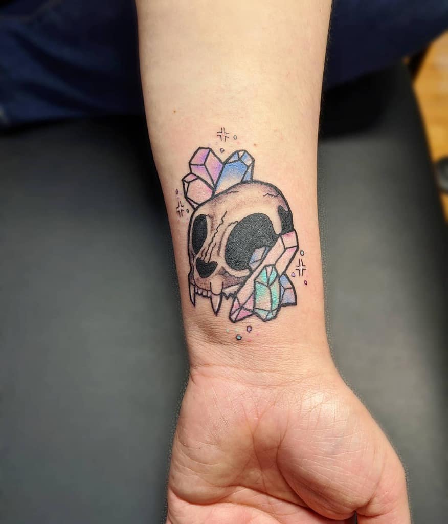Tattoo of a stylized cat skull adorned with colorful crystals on a wrist