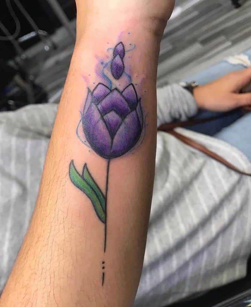 A vibrant purple tulip tattoo on the forearm with soft shading and colorful accents, including a flowing stem and decorative details.