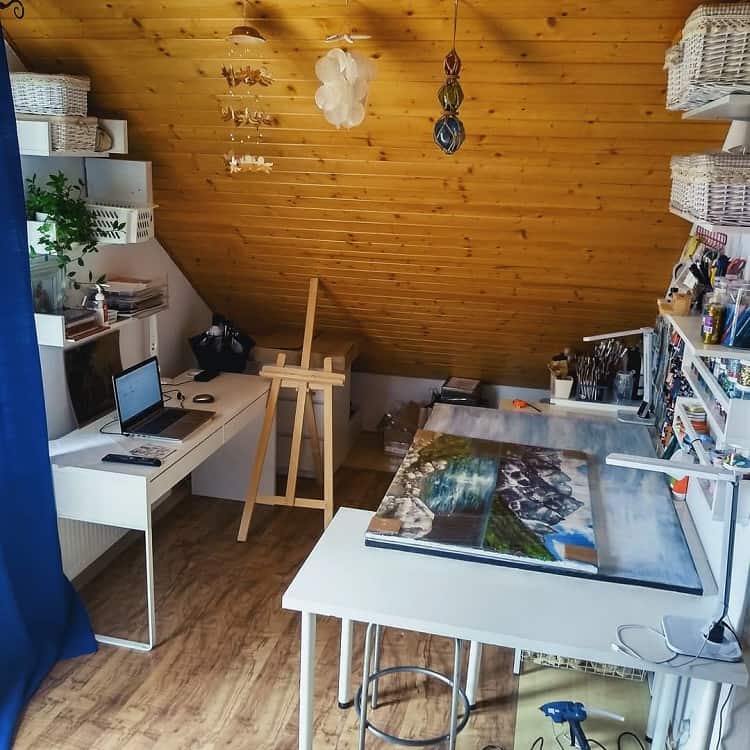 Cozy attic art studio with a slanted wooden ceiling, easel, laptop, and art supplies organized on shelves