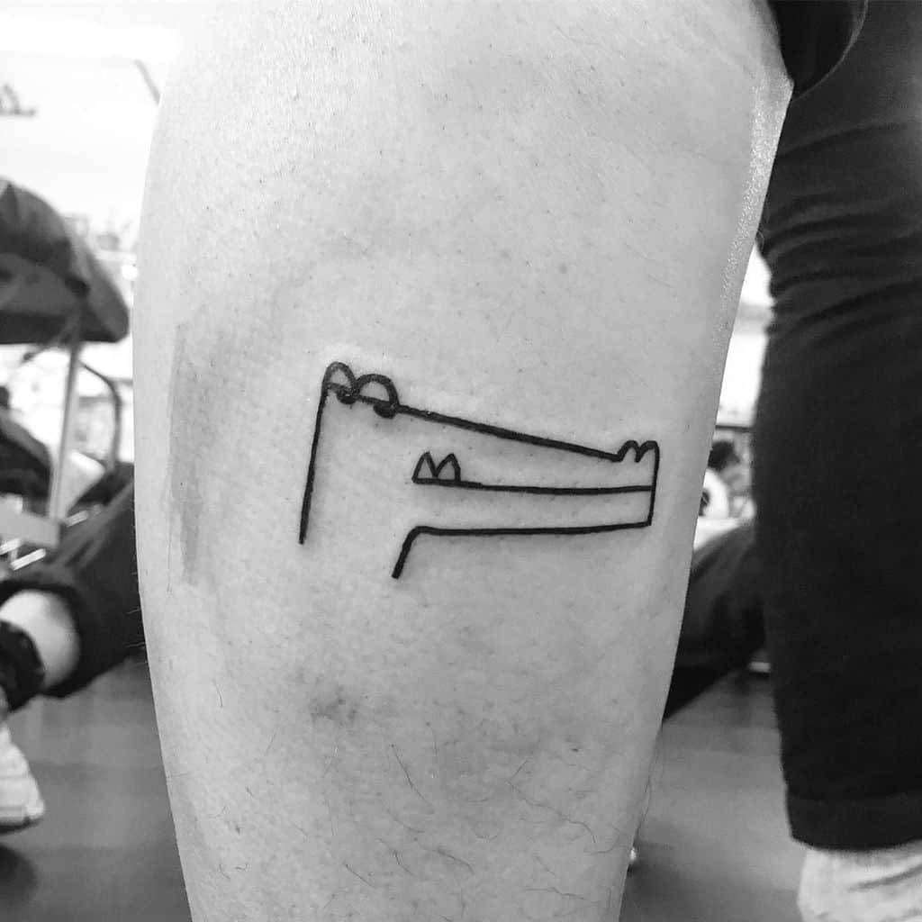 A minimalist black ink tattoo of a crocodile drawn with simple lines, placed on the leg for a playful and abstract look.
