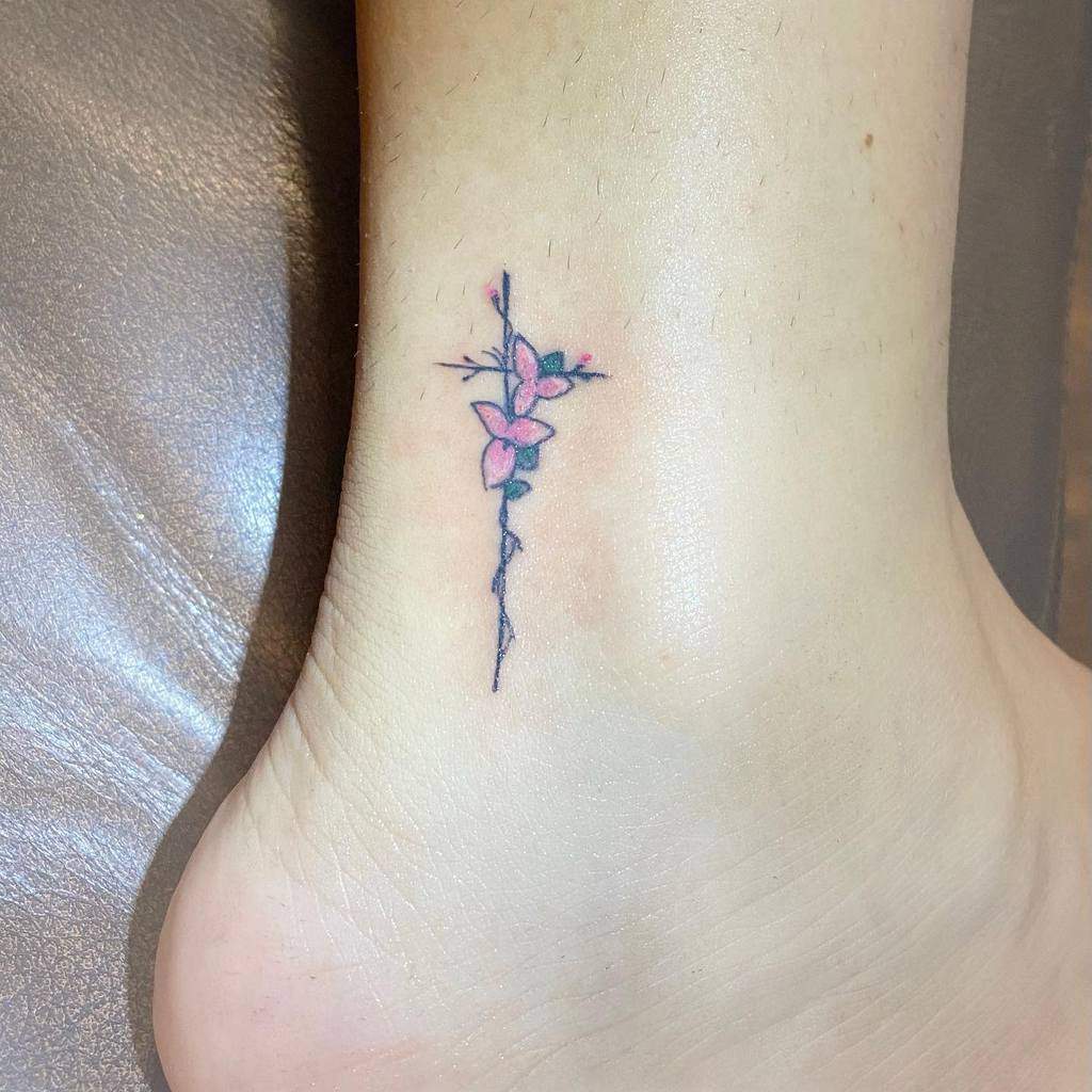 A small tattoo near the ankle featuring a delicate branch with pink flowers, intricately forming the shape of a cross