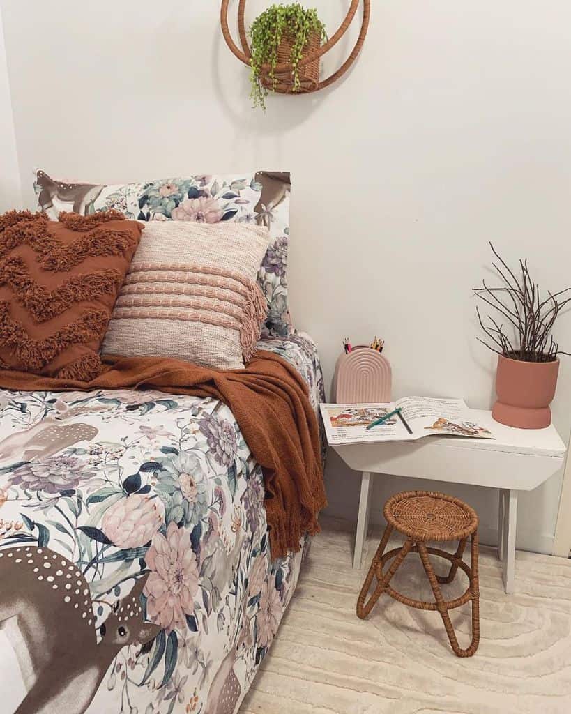 Cozy bedroom corner with a floral bedspread, pillows, small table, stool, potted plant, and a hanging plant