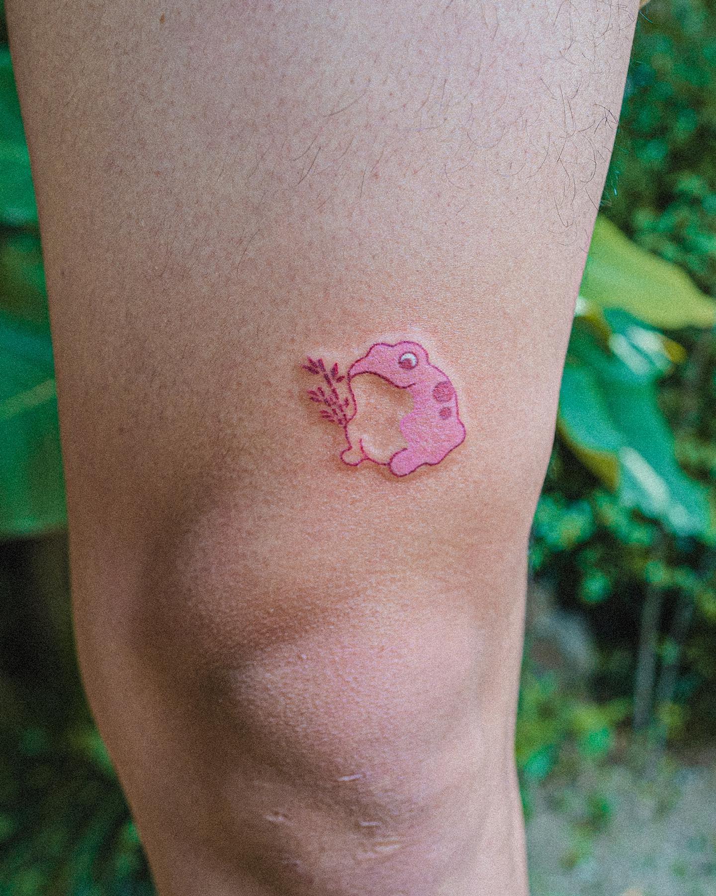 Minimalist pink frog tattoo holding a sprig of leaves
