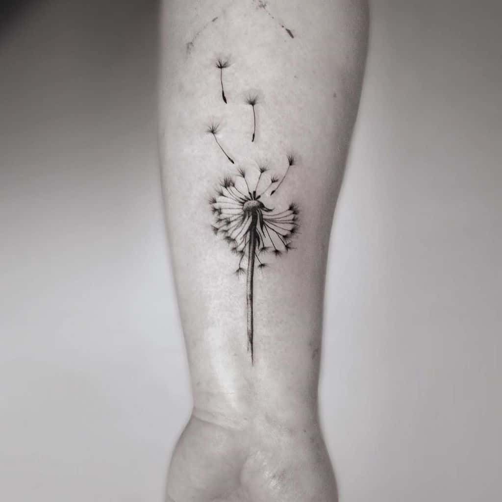 Black ink dandelion tattoo on the forearm with seeds drifting away, symbolizing wishes, change, and letting go.