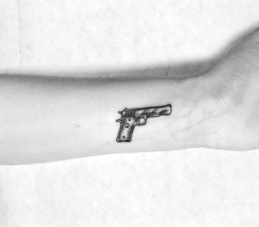 Minimalist black and gray tattoo of a small 1911 pistol on the wrist, featuring fine line work and subtle shading for a simple yet bold design.