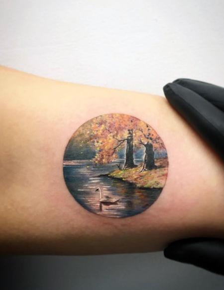 Small circular tattoo of a serene autumn lakeside scene with golden trees, two figures walking, and a swan gliding across the water.