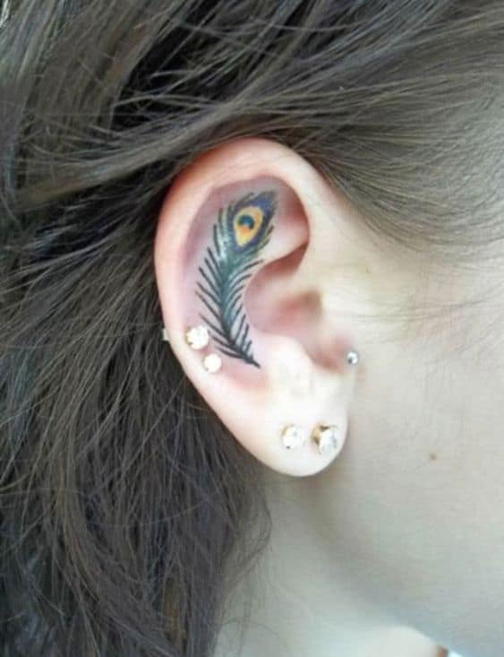 Small and intricate peacock feather tattoo inside the ear, featuring delicate green and yellow hues with fine detailing for a subtle look.