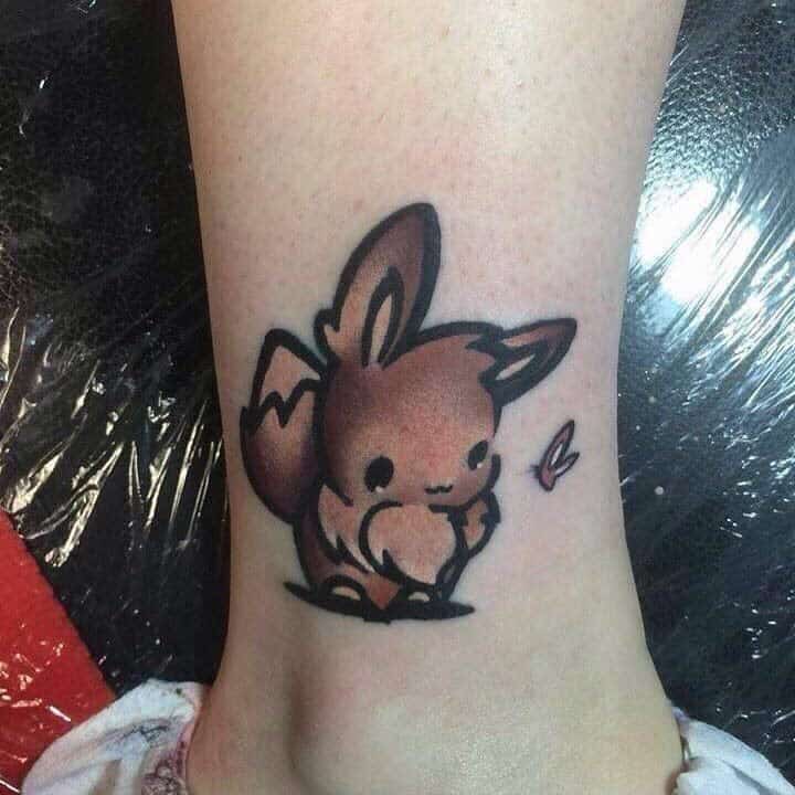 Adorable chibi-style Eevee tattoo with soft brown tones and a tiny pink butterfly accent near the foot
