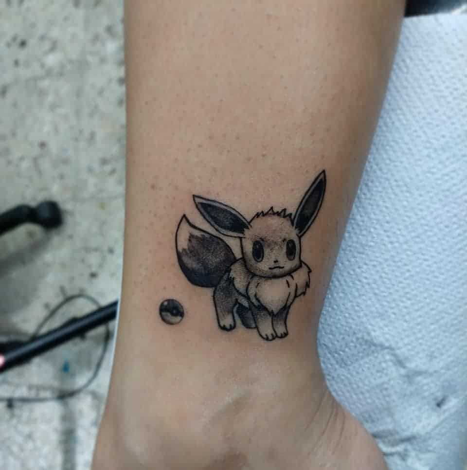 Black and white tattoo of Eevee from Pokémon on a person's arm, with a small Pokéball next to it