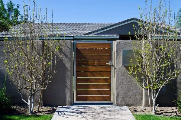 Wooden gate set in a stone wall, flanked by two leafless trees, with a house and clear blue sky in the background