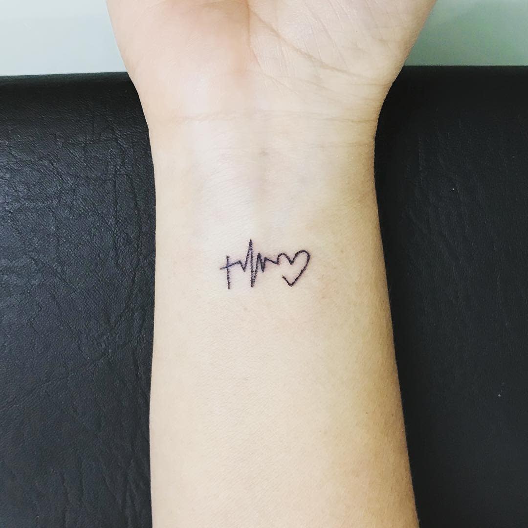Simple wrist tattoo featuring a small cross, heartbeat line, and heart symbol