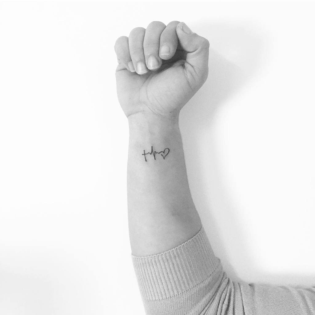Minimalist tattoo on the wrist featuring a small cross, heartbeat line, and heart symbol