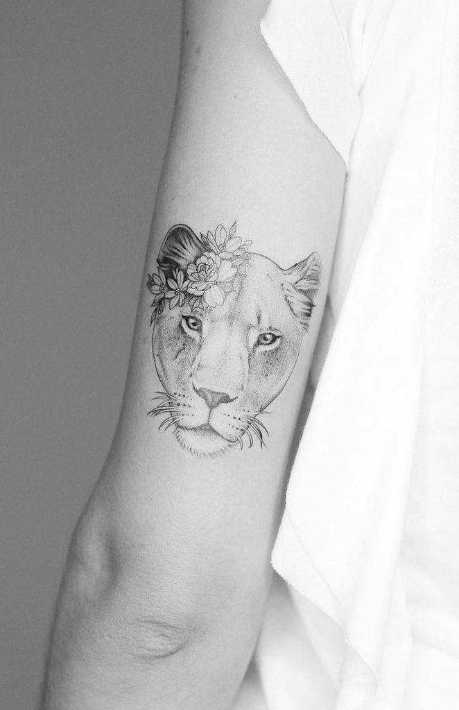 Elegant black and grey lioness tattoo on the upper arm, featuring fine details and a floral crown for a feminine yet powerful look.